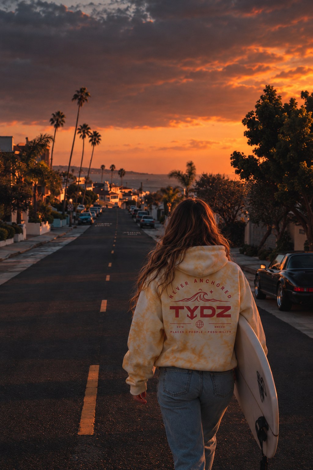 Woman wearing a TYDZ hoodie and carrying a surfboard on a coastal road at sunset